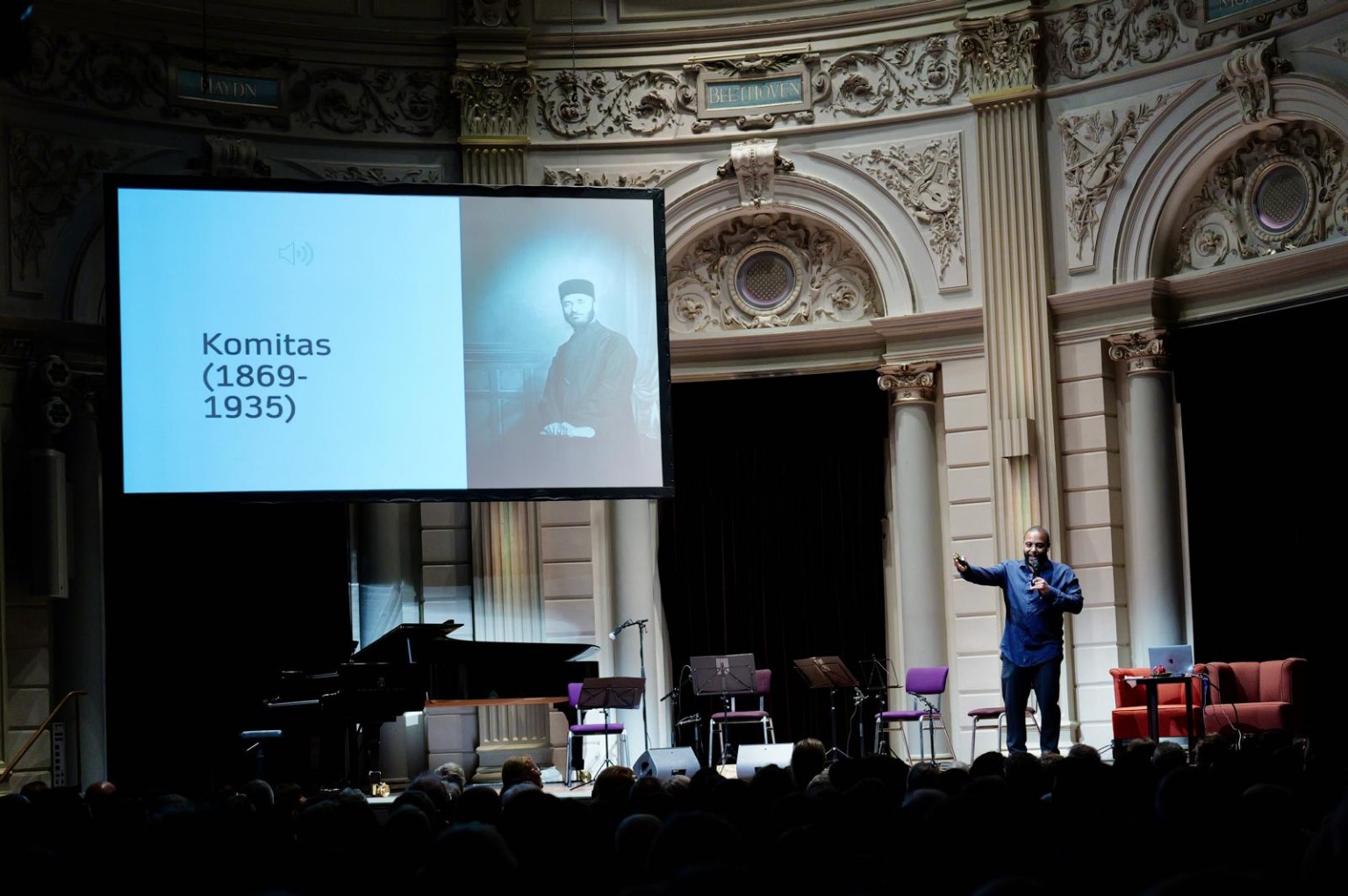 Orville Breeveld speaking on the Kleine Zaal stage beside a projected photograph of Gomidas.